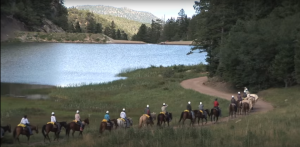 Horseback riders follow a trail alongside a lake with mountains in the background
